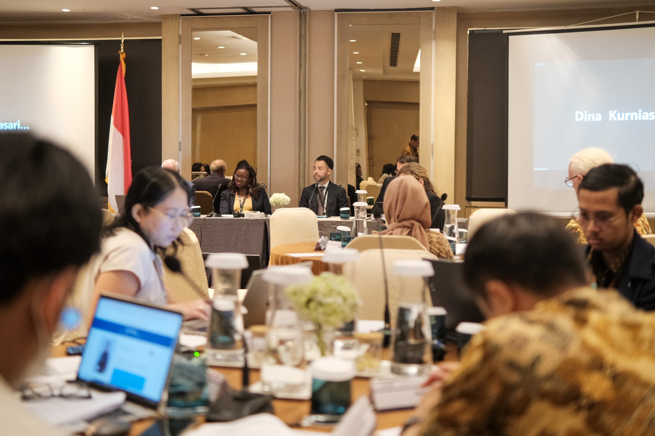 This is a photo with panelists of the program in the background. The foreground includes participants on their laptops with microphones in front of them, paying attention to the panel on a round table.