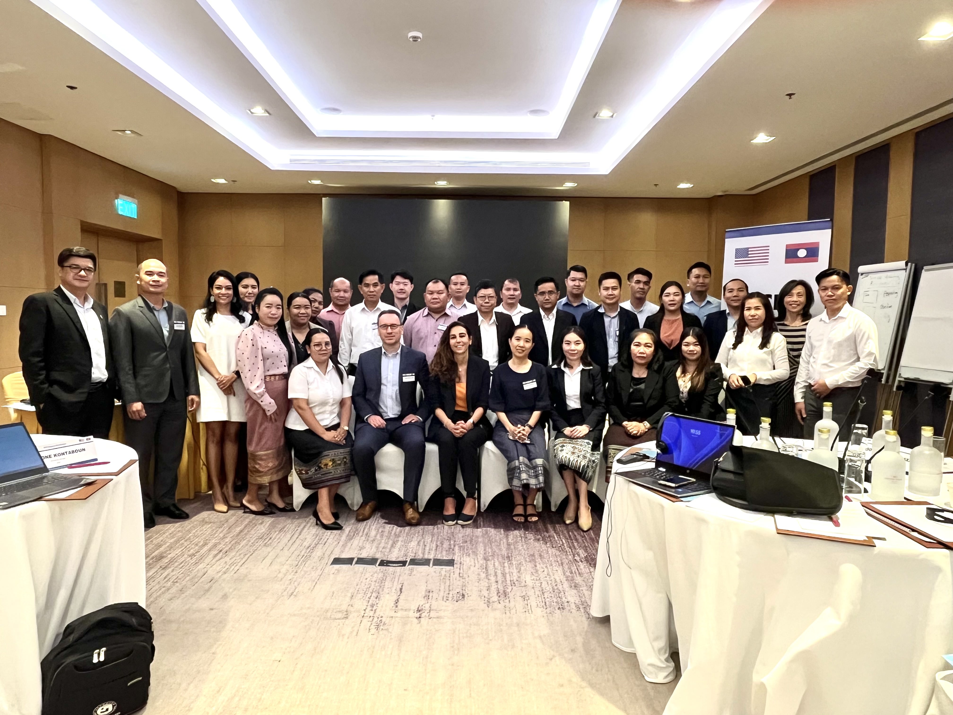 This is a group photo of participants in the Laos PPP Dispute Resolution workshop. In the foreground there are women sitting on chairs, and in the back there are 20 or so participants standing. All participants are smiling.