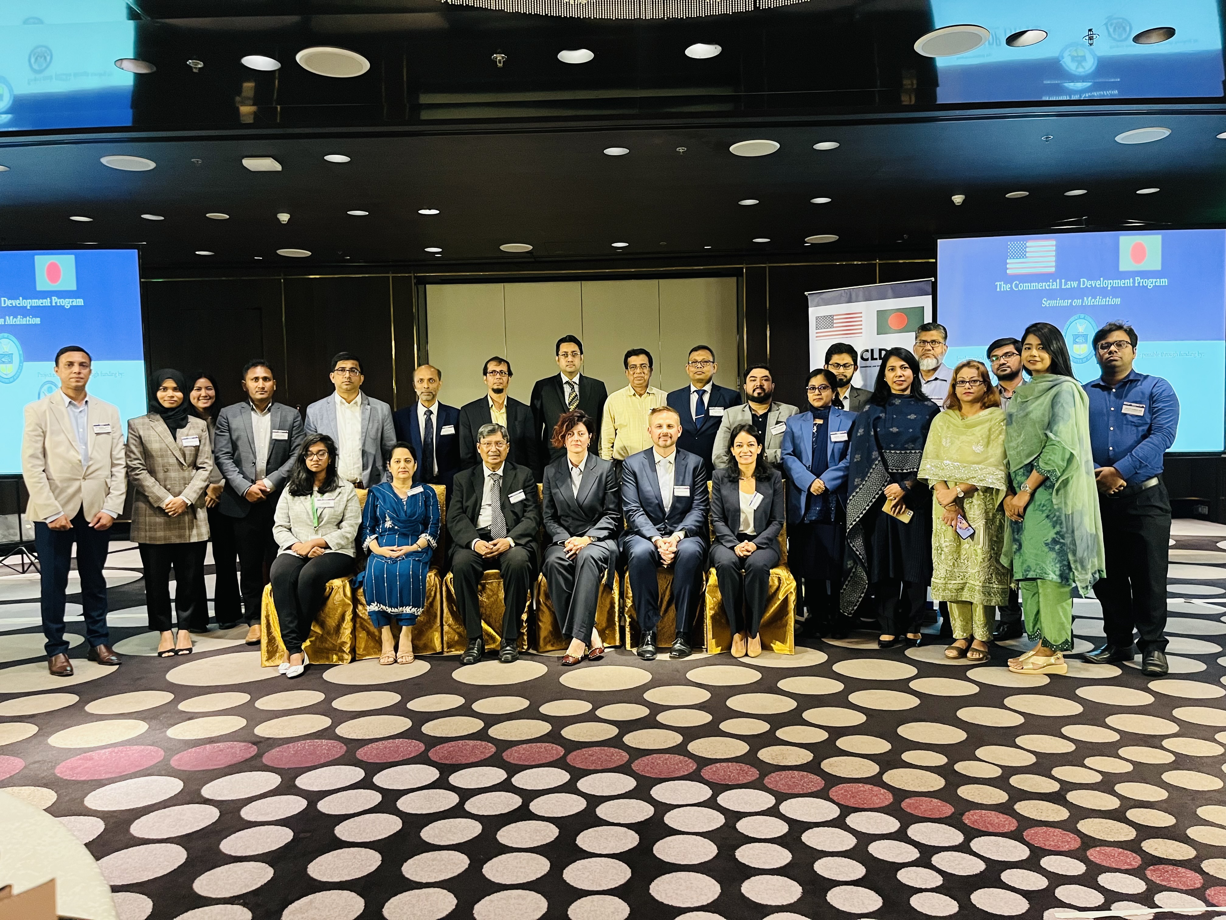 Group photo of participants in a hotel conference room bisected by two screens titled &quot;Mediation Workshop&quot;