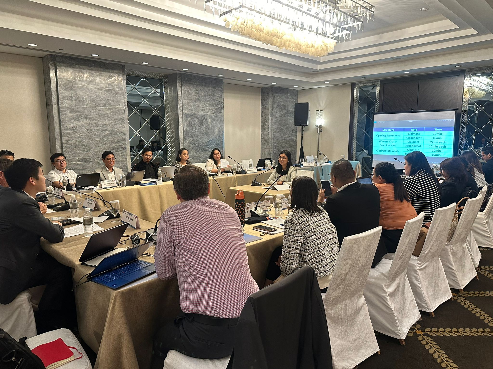 Participants sitting in a U-shape setup in a conference room with one participant in the center giving a witness statement