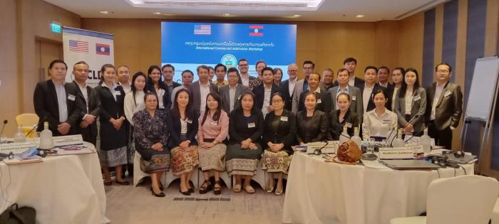 This is a group photo of participants in the Laos International Commercial Arbitration workshop. In the foreground there are women sitting on chairs, and in the back there are 20 or so participants standing. All participants are smiling.
