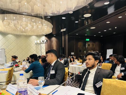 This is a zoom in photo of two male participants from the Bangladesh ADR workshop. The men are seated at a white table with golden clothes chairs. There are water bottles, glasses, nametags, and notebooks in front of them.